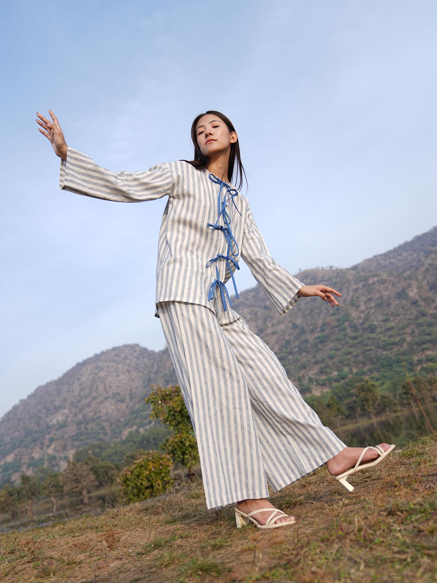 Woman wearing the Sutra Bow Stripe co-ord — an open-front striped cotton tunic tied with blue fabric ties over wide-leg trousers