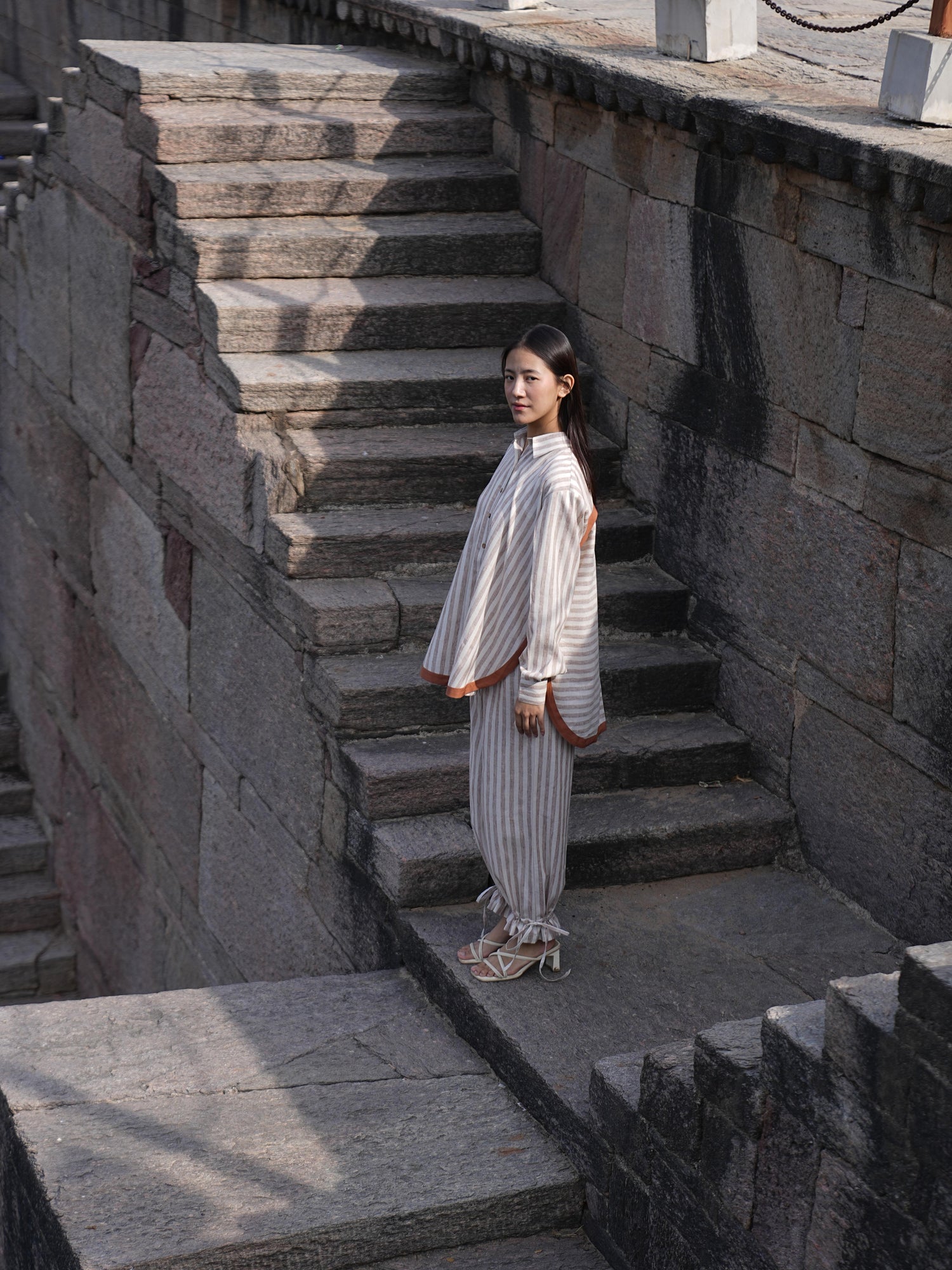 Woman wearing the Sutra Arc Stripe co-ord — a striped cotton button-down shirt with matching ruffled-hem trousers